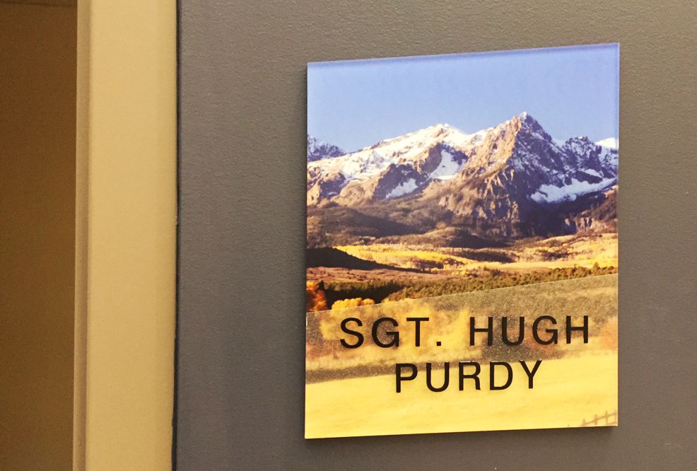 A wall plaque with a scenic background of snow-capped mountains and autumnal trees has the text "Sgt. Hugh Purdy" printed on it. The plaque is mounted on a gray wall next to a beige door frame.