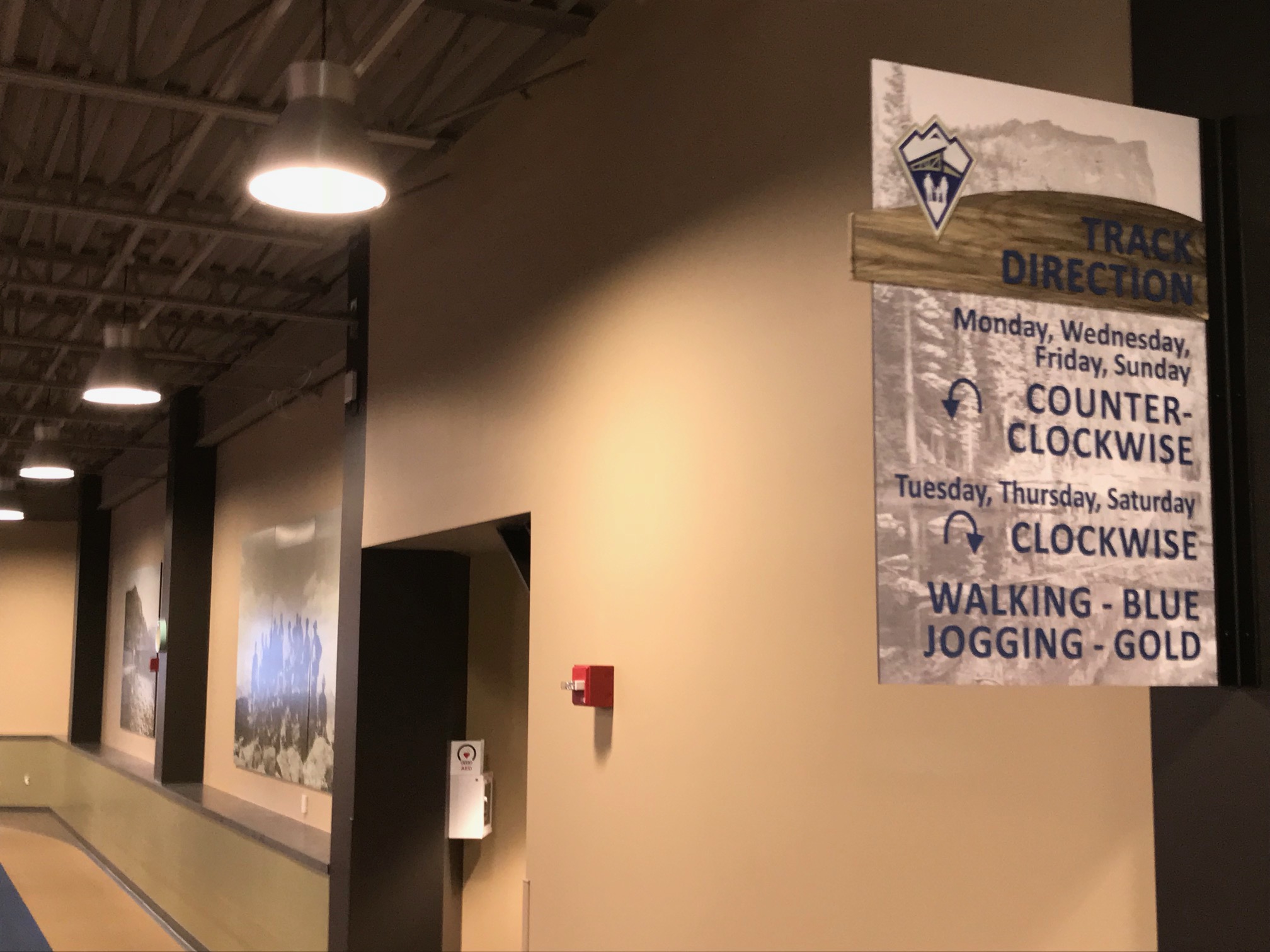 Indoor track with beige walls and ceiling lights. Sign on wall reads "Track Direction" with alternating instructions for clockwise and counterclockwise directions, and color codes for walking (blue) and jogging (gold) lanes. Various photographs are displayed on the wall.