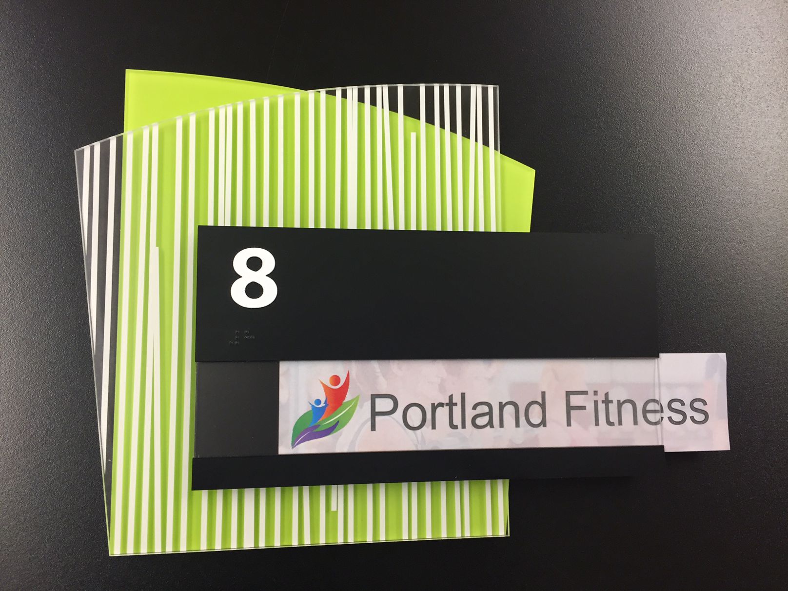 A black rectangular sign with the number "8" in white at the top. Below it, there is a colorful logo of three human figures and the text "Portland Fitness" on a white background, partially slid out to show that the sign is editable. The sign is set against a background of vertically striped green and white paper.