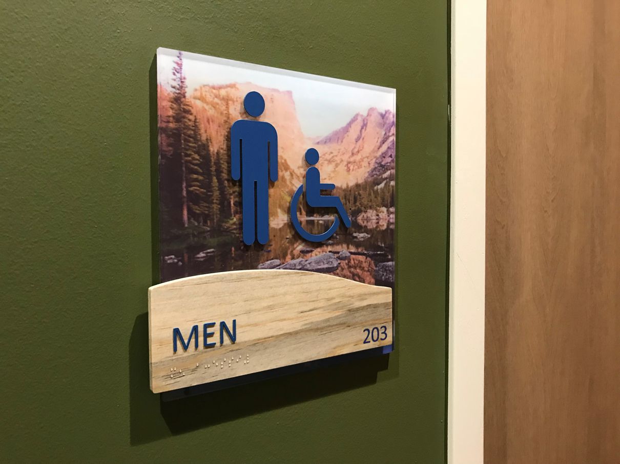 A restroom sign on a wall indicating a men's restroom, featuring symbols for men and accessibility for people with disabilities. The background includes a landscape image, and the sign is labeled "MEN" with the number "203" at the bottom.