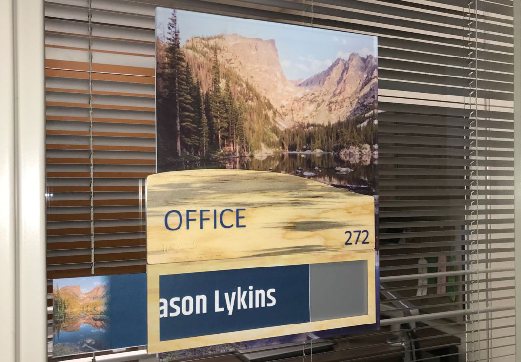 A glass door with blinds features a sign displaying a nature scene and the text "OFFICE 272 Jason Lykins." The office interior is visible through the window.