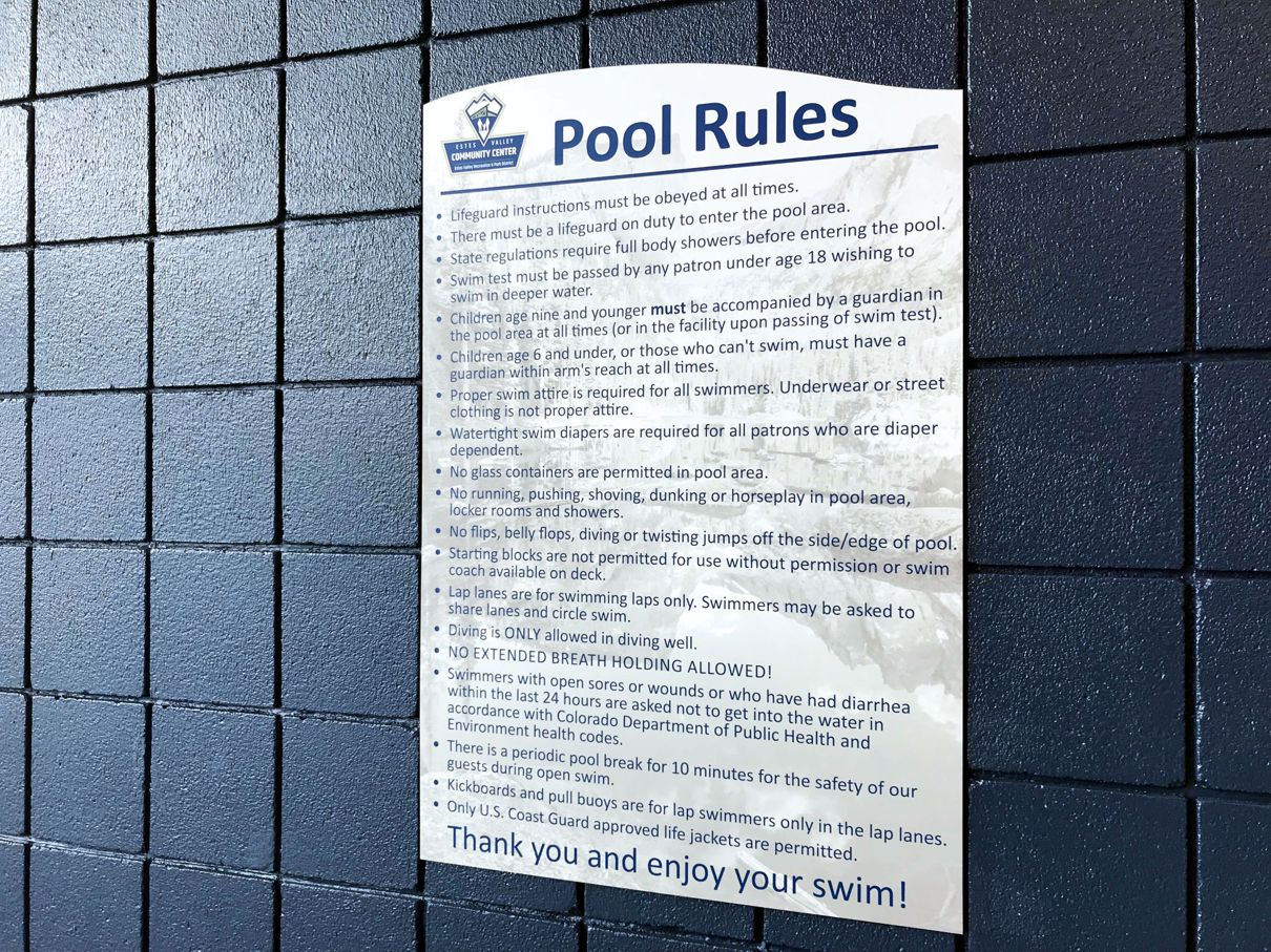 A sign posted on a tiled wall lists the pool rules, including requirements for lifeguard presence, swim attire, activity regulations, and hygiene protocols. The sign encourages reading all rules thoroughly and concludes with a reminder to enjoy the swim.