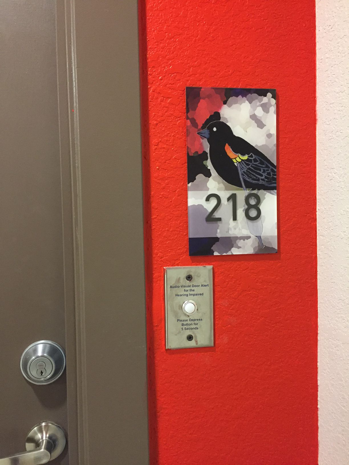 A door with room number "218" sign beside it, featuring a colorful bird illustration. Below the sign, there's a silver intercom panel with a white button labeled "Press to Open" and "Assistance/Presione Para Ayuda." The wall is painted bright red.