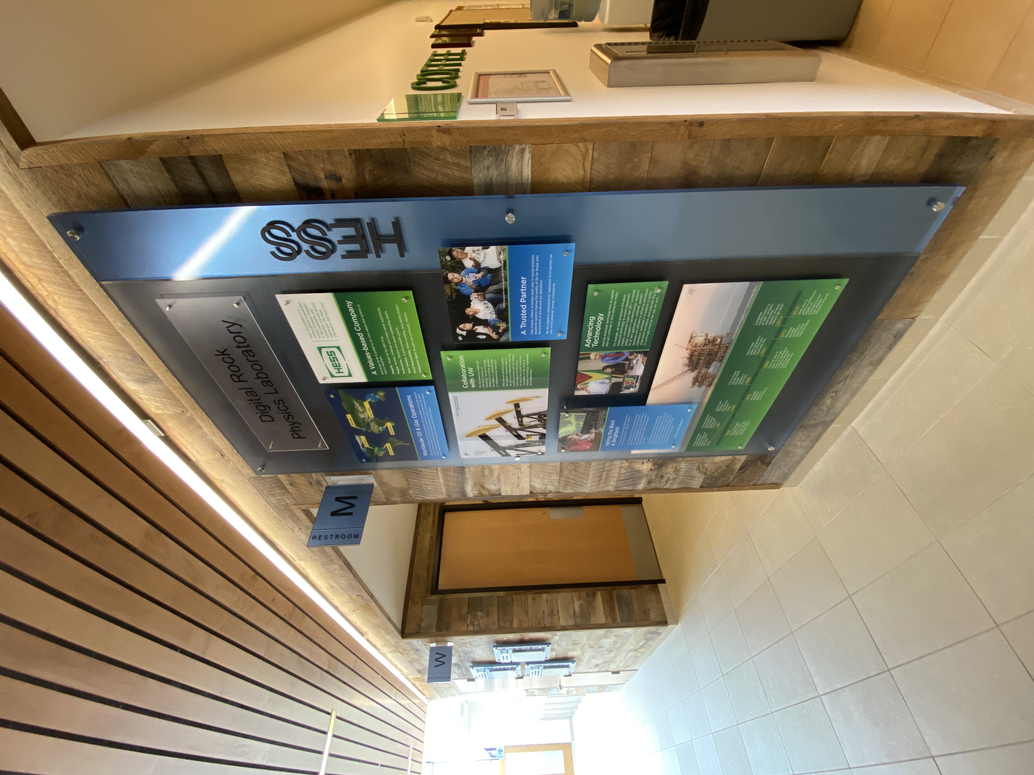 A hallway with a bulletin board featuring the Digital Rock Physics Laboratory at the HESS building. The board showcases various informational posters and images. To the left, there are wooden doors labeled with a 'W.' The ceiling has wooden paneling, and there's a coffee machine visible to the right.