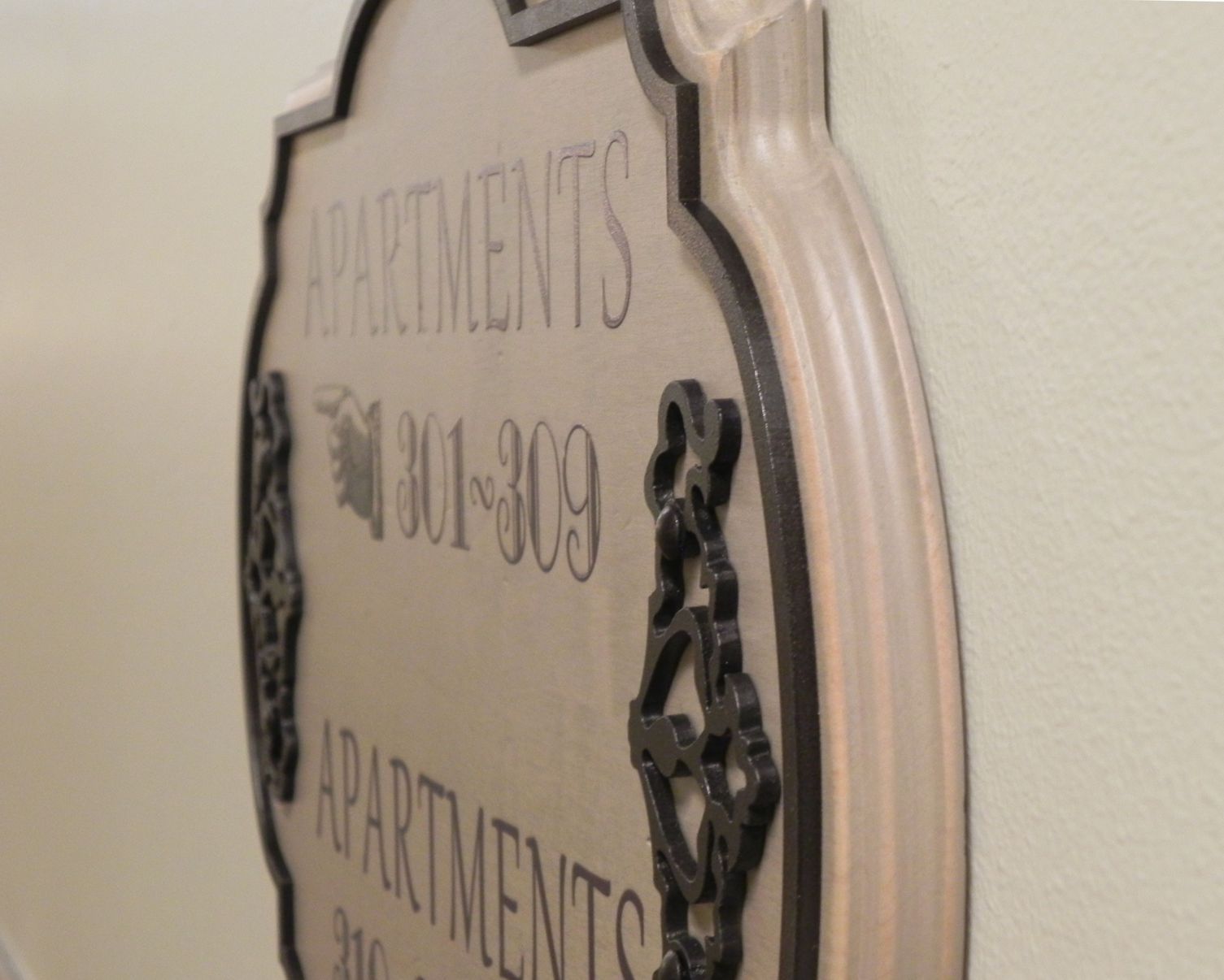 Side view of a decorative sign mounted on a wall. The sign reads "APARTMENTS 301-309" in embossed letters. The sign has an ornate design with intricate black detailing along its edges. The wall behind the sign is off-white.