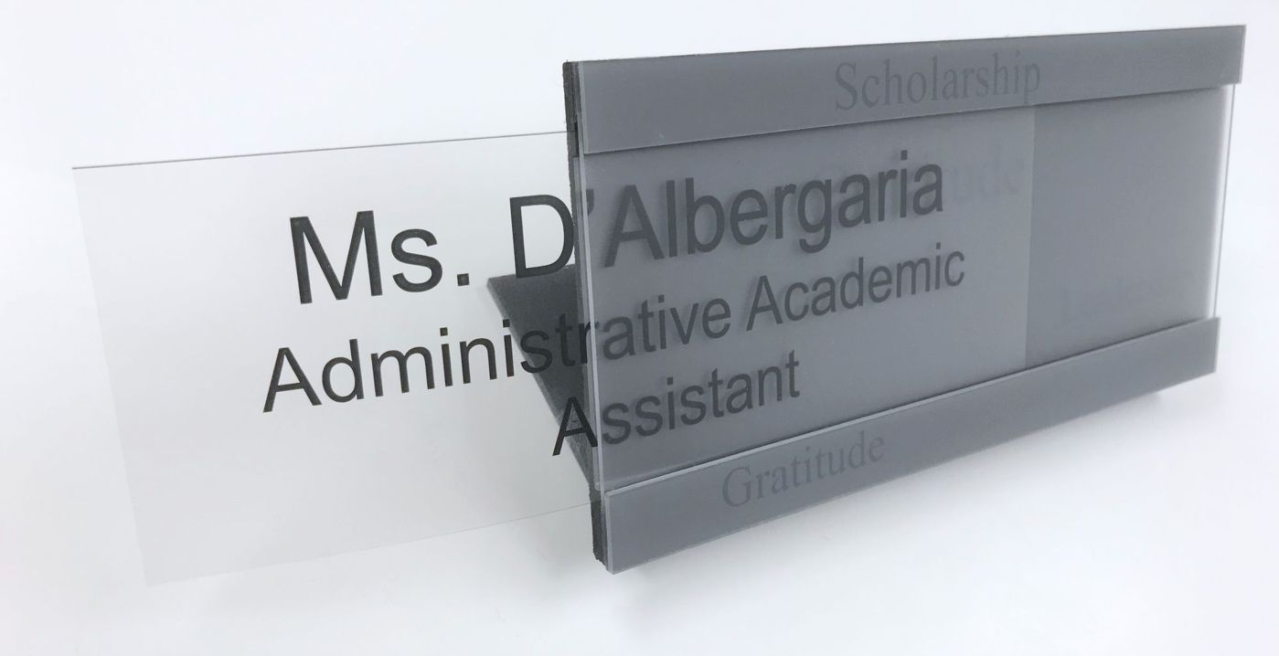 A gray desk nameplate with black lettering reads "Ms. D'Albergaria, Administrative Academic Assistant." The word "Scholarship" is engraved at the top, and "Gratitude" is engraved at the bottom.  The clear plate with text is half slid out of the holder.
