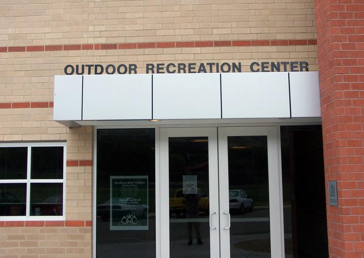 Entrance of the Outdoor Recreation Center with glass doors and a white awning above them. The building is made of beige and red brick. There is a sign on the door with icons indicating bicycle-friendly facilities.