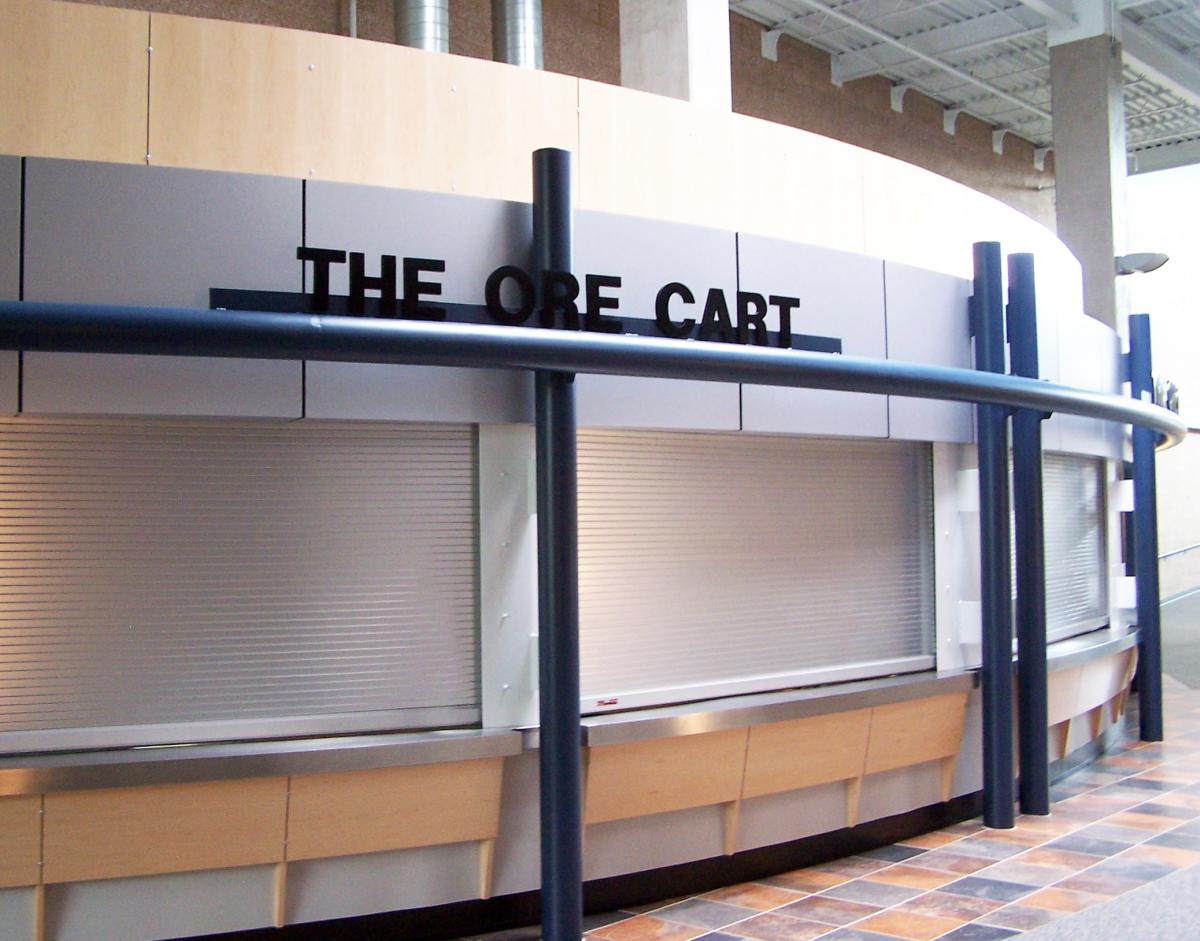 A modern storefront with a sign reading "THE ORE CART." The structure features sleek blue and silver designs, with a partially visible roller shutter door. 