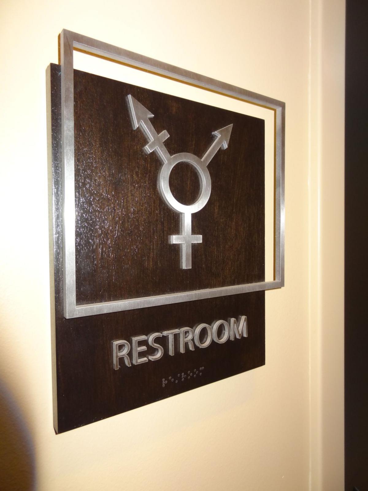 A sign on a wall denotes a restroom with a symbol combining male, female, and non-binary gender symbols. The word "Restroom" is written below the symbol along with braille text.