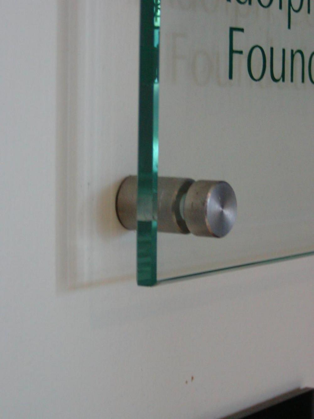 A clear glass plaque mounted on a wall, held by four metal fasteners. This is a closeup of one of the corner fasteners.