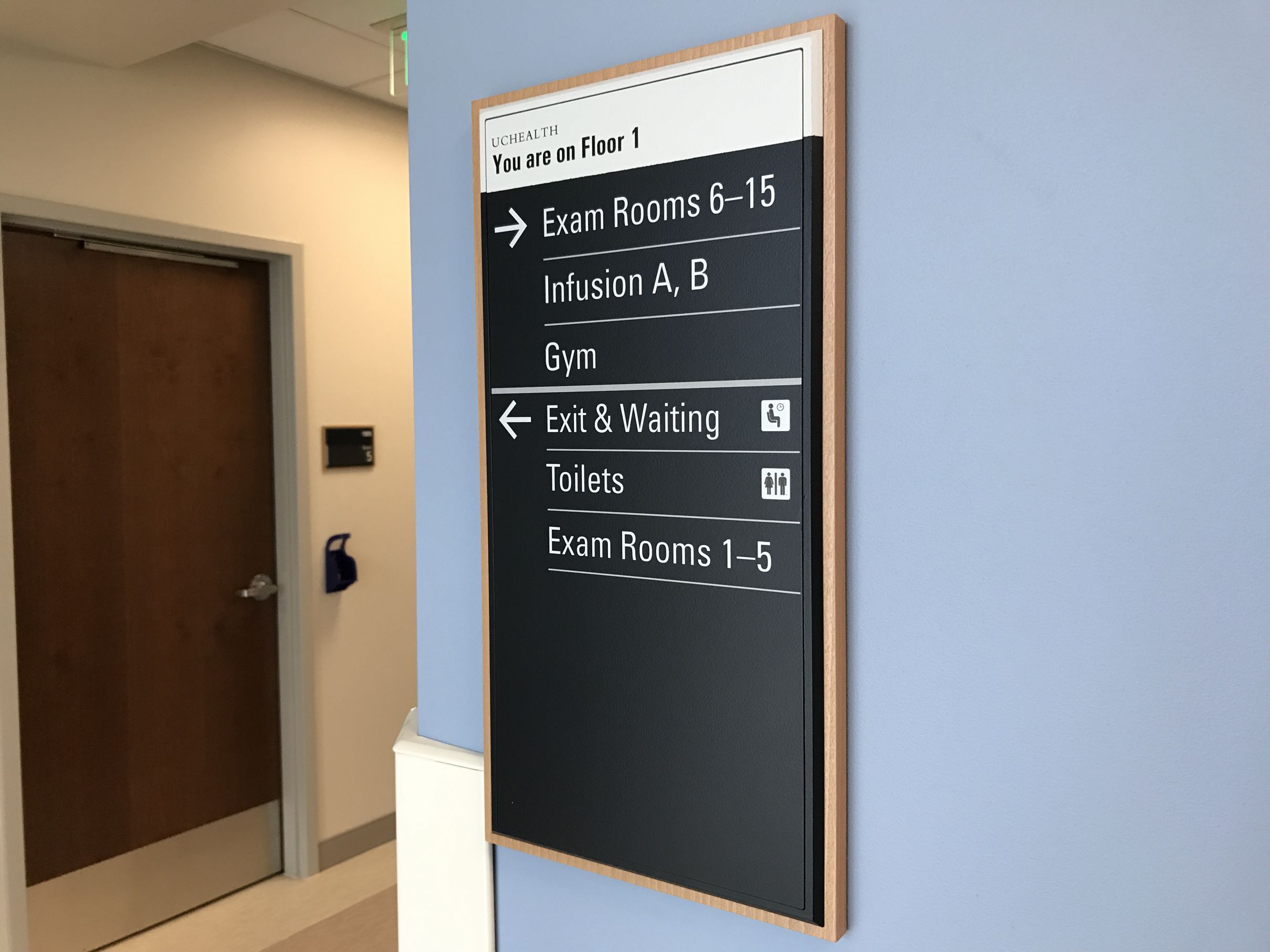 A directional sign inside a healthcare facility indicates that on Floor 1, Exam Rooms 6-15, Infusion A and B, and the Gym are to the right. The Exit & Waiting area, Toilets, and Exam Rooms 1-5 are to the left. A wooden door is visible to the left of the sign.