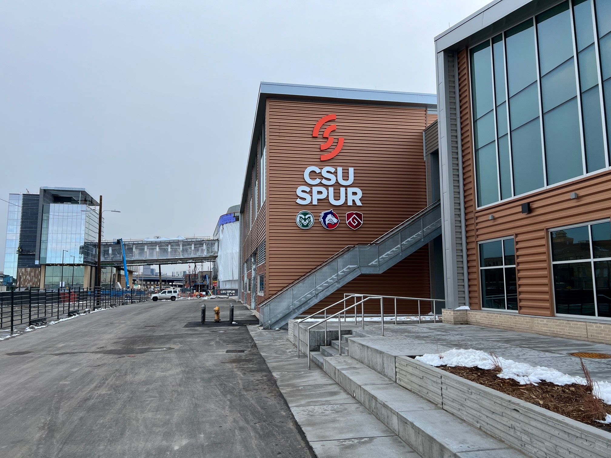 A modern building with large "CSU SPUR" sign and three illuminated logos on its facade. Snow covers the ground and an overpass bridge is visible.
