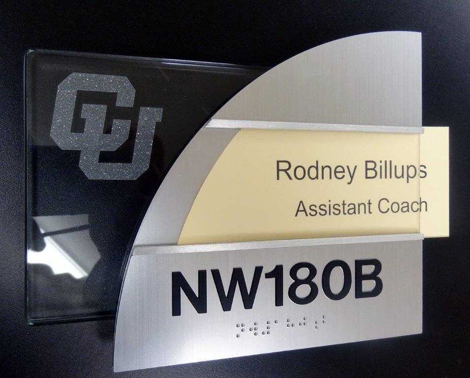 A nameplate with an angular design showing the text "Rodney Billups, Assistant Coach," and the room number "NW180B" in bold black letters. The nameplate also features the CU logo and Braille markings. The background is black.