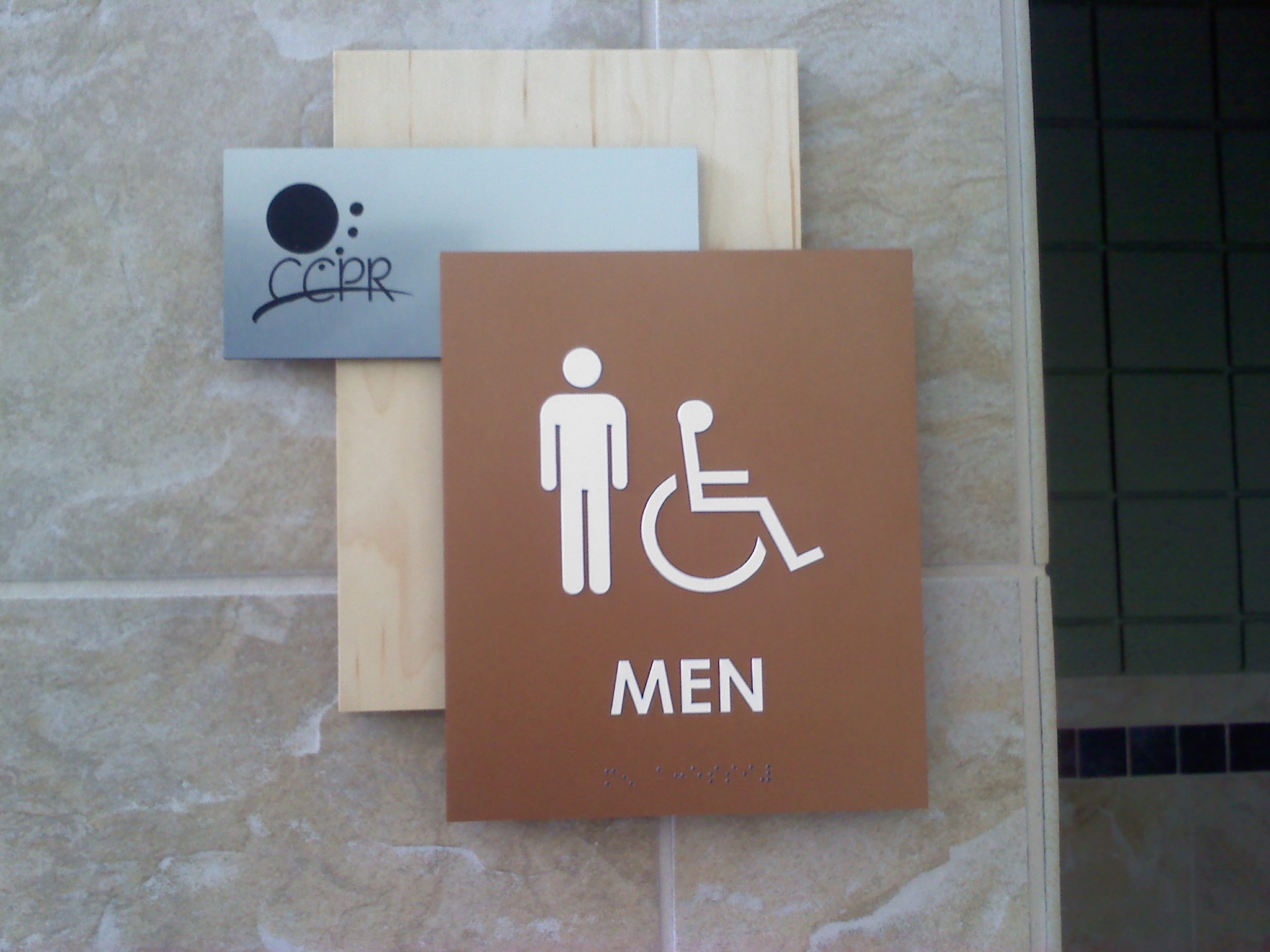 A restroom sign on a tiled wall displays symbols for men and accessibility for individuals with disabilities. The word "MEN" is written below the symbols. A smaller sign above the restroom sign has the text "CCPR" and an abstract design.
