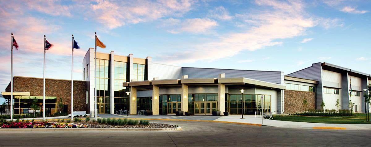 A modern commercial building with a glass facade is seen against a backdrop of a partly cloudy sky. Four flagpoles with various flags stand to the left of the entrance. There is a landscaped area with flowers along the driveway leading to the entrance.