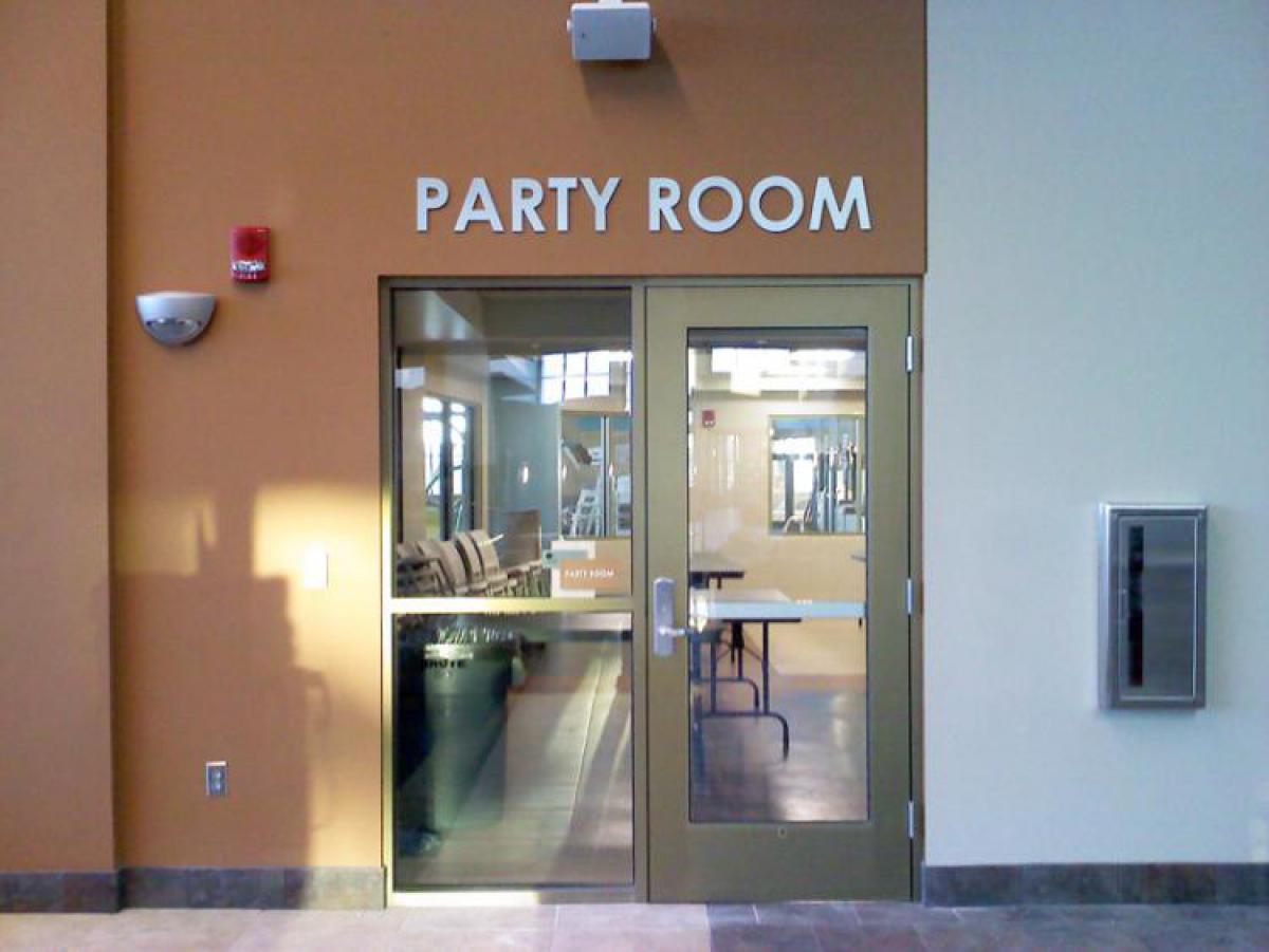 Entrance to a room labeled "Party Room" with glass double doors. The doors provide a view of a few tables and chairs inside. The surrounding area has beige walls, a wall-mounted light, a fire alarm, and an empty wall-mounted metal box on the right.