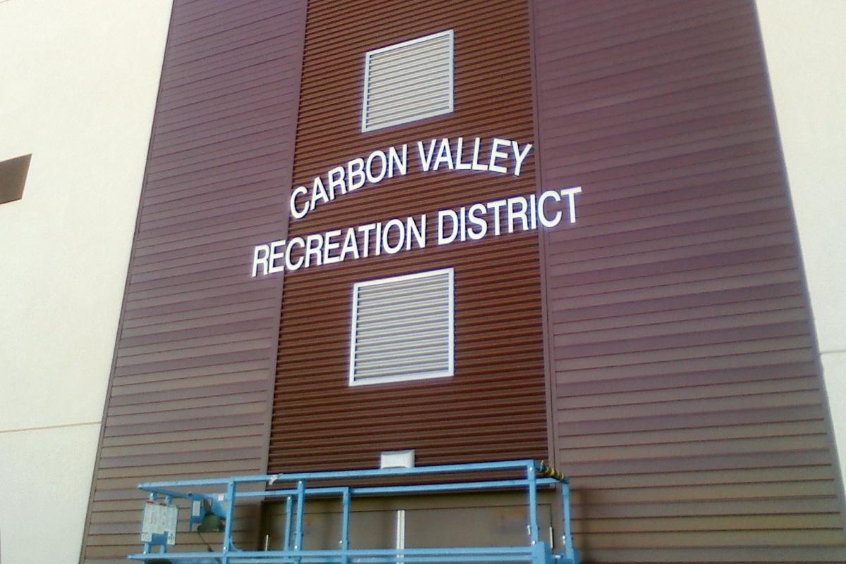 The image shows the exterior wall of a building with the words "Carbon Valley Recreation District" displayed in large, white letters. The wall is brown with vertical panels, and there is some blue scaffolding near the base of the wall.