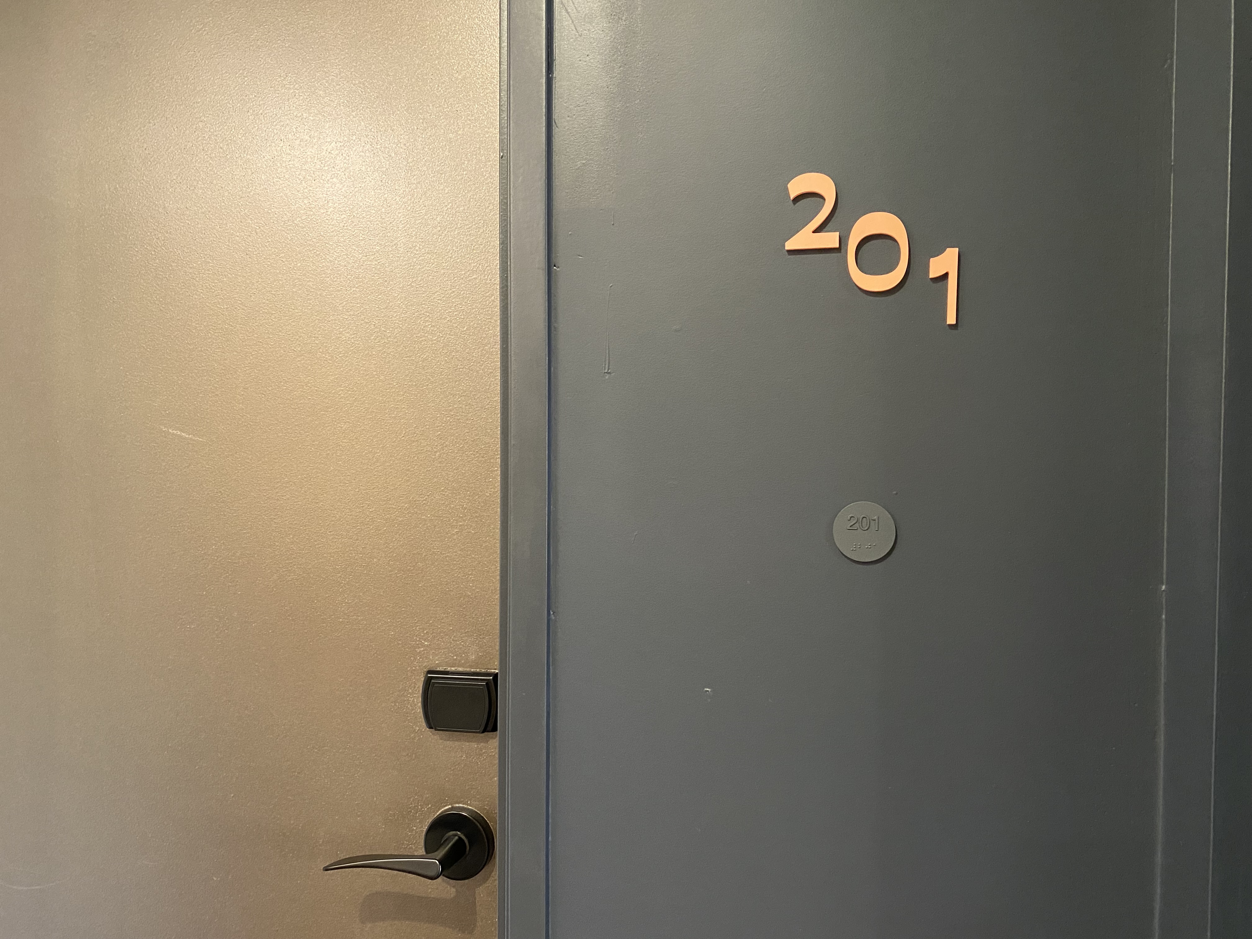 A close-up view of a closed door painted in a beige color with a dark handle and an electronic lock. The adjacent wall is dark, featuring the room number "201" displayed in copper-colored numbers.  A smaller circular button reads "201" in raised numbers and braille.