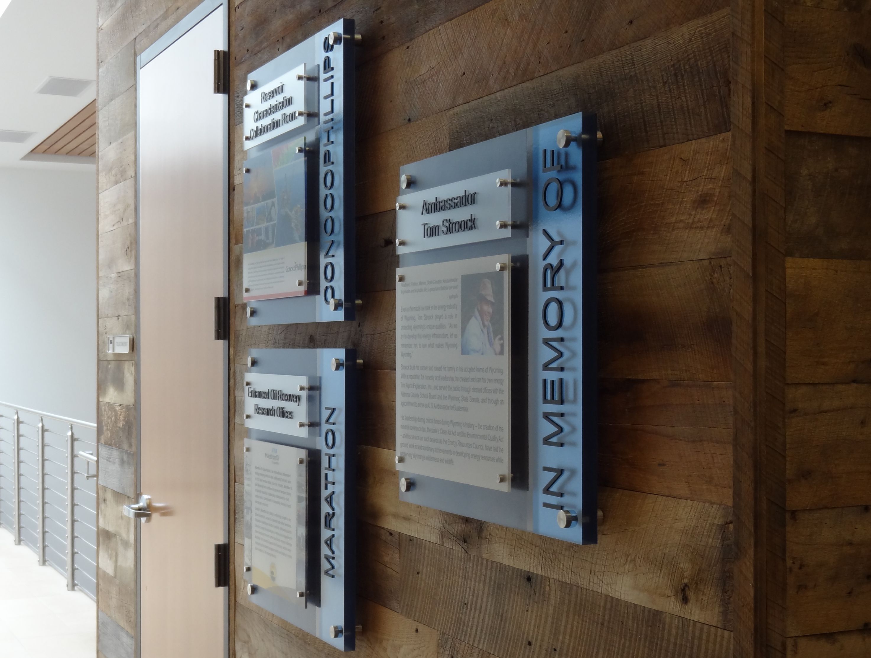 Wood-paneled wall adorned with three rectangular plaques with text and images. Titles on the plaques include "Ambassador Tom Simock." Vertical blue text along the edge reads "IN MEMORY OF". Adjacent open door and metal railing on the left.
