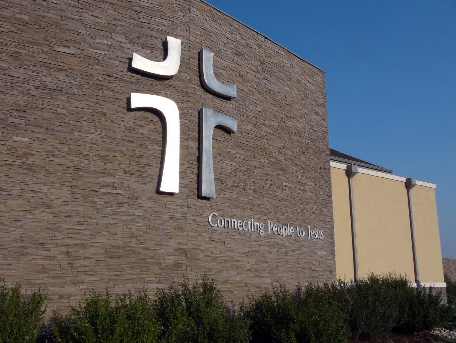A modern church building with a large, stylized cross on its brick wall. Below the cross, the phrase "Connecting People to Jesus" is visible. The church is surrounded by neatly trimmed greenery and set against a clear blue sky.