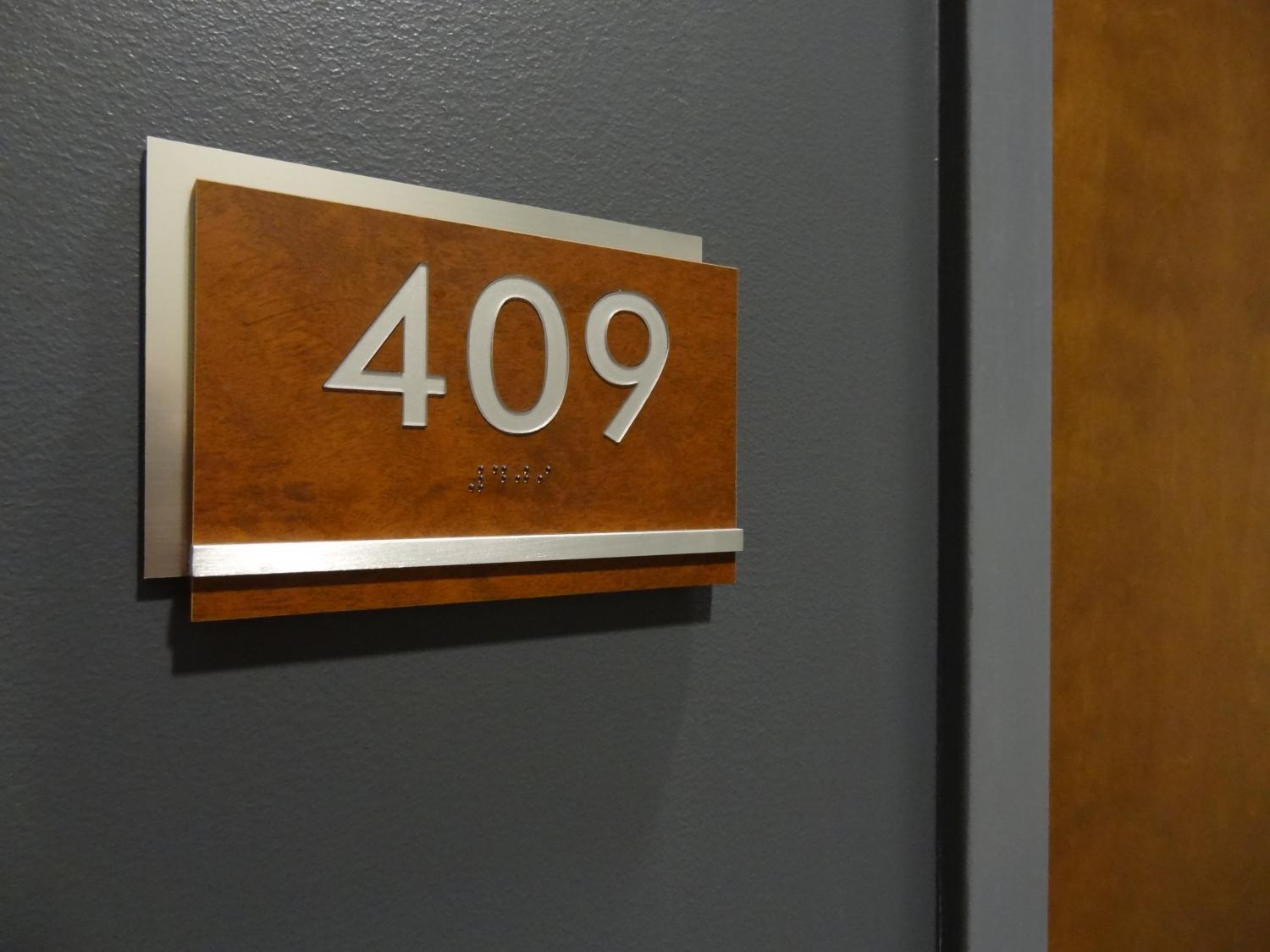 A wall-mounted sign displaying the room number "409" in silver numerals on a wooden background. Beneath the numerals, there are tactile braille characters. The sign is on a gray wall next to a wooden door.
