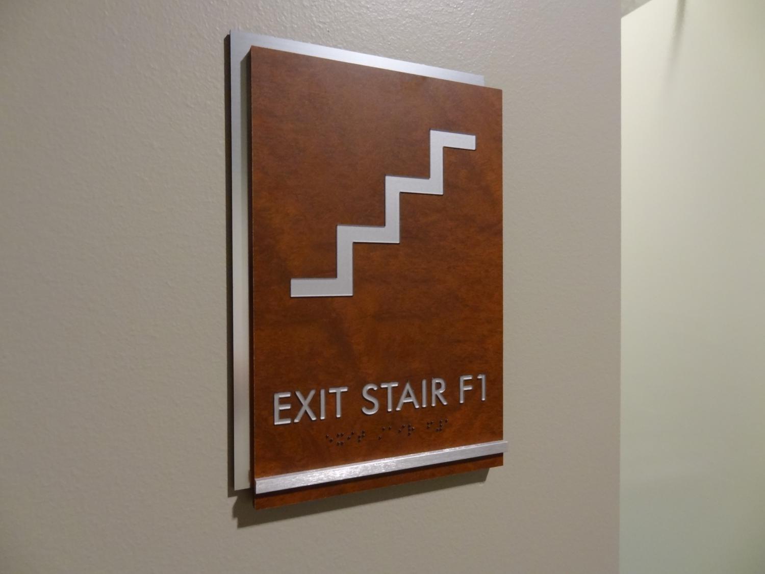 A wall-mounted sign with a wooden finish displays a white graphic of a staircase above the text "EXIT STAIR F1." Below the text, there are raised Braille characters.