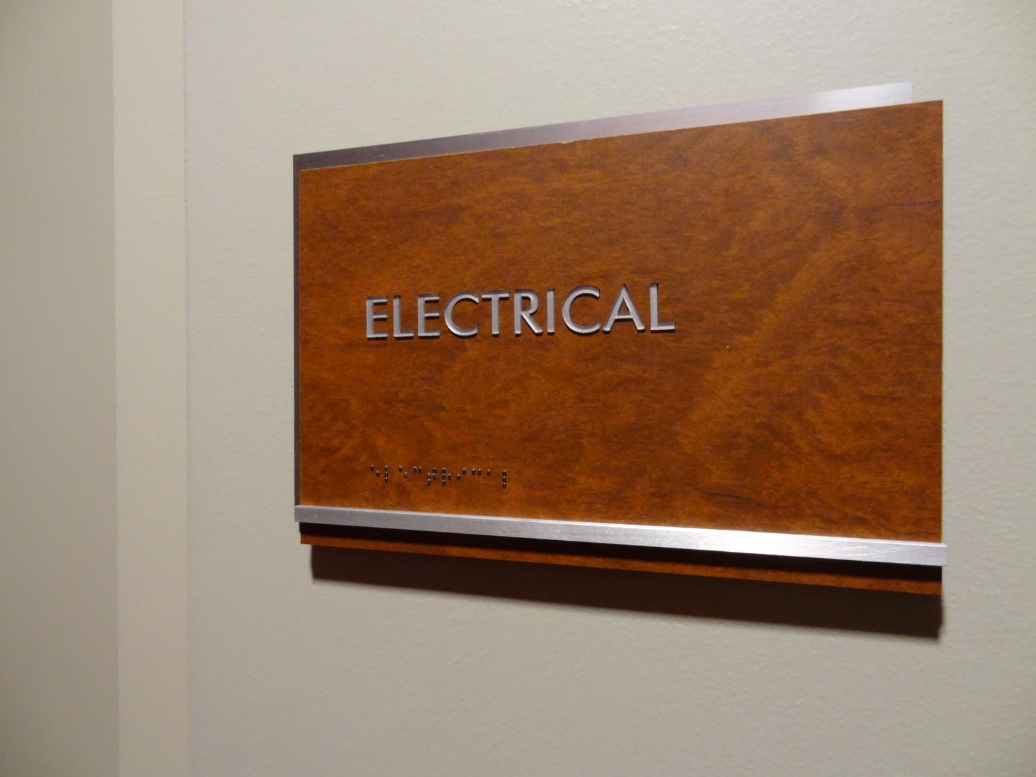 A wooden door sign reads "ELECTRICAL" in uppercase silver letters, mounted on a beige wall. Braille text is displayed beneath the main text.