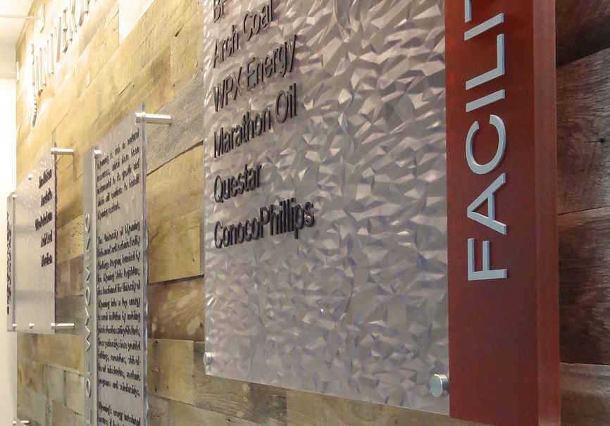 A wall display features large plaques with text, some of which list names such as BP, Arch Coal, WPI Energy, and ConocoPhillips. The background consists of wooden panels, and a red vertical plaque on the right has "FACILITY" written on it.