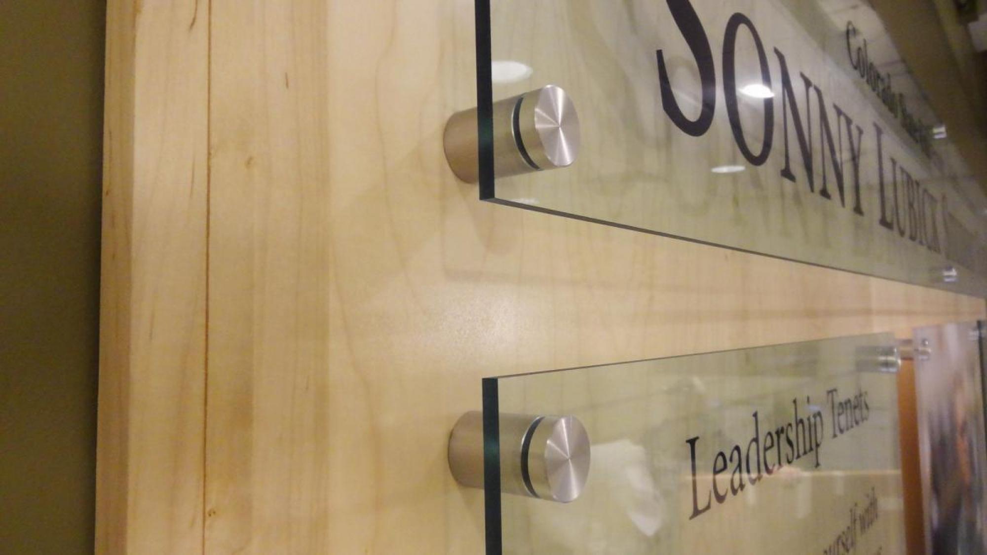 A close-up side view of glass plaques mounted on a wooden surface with metal spacers. The plaques contain text, but only partial words are visible, such as "Sonny" and "Leadership Tenets." The background is a light-colored wooden wall.