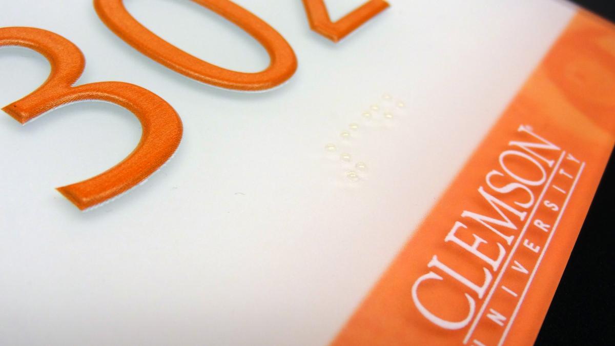A sign reads "302" in large orange numbers on a white background. Below the numbers, there is a marbled orange strip with "Clemson University" written in white uppercase letters.  This is a closeup that also shows the braille text beneath the numbers.