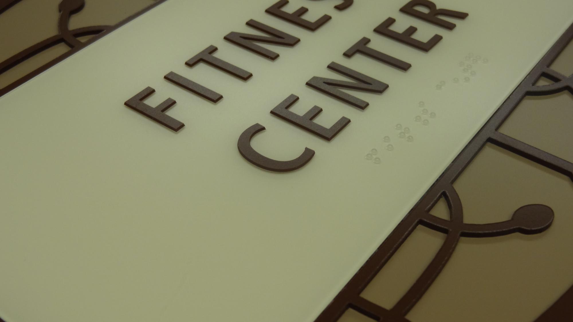 Close-up of a sign that reads "Fitness Center" in raised, dark brown letters with braille directly below the text. The background is a light beige color, and the sign is bordered by an intricate, decorative pattern.