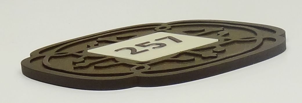 A close-up photograph of an oval-shaped, carved wooden plaque with an intricate pattern and the number "257" displayed prominently in the center on a white background. The plaque is resting on a flat, neutral-colored surface.