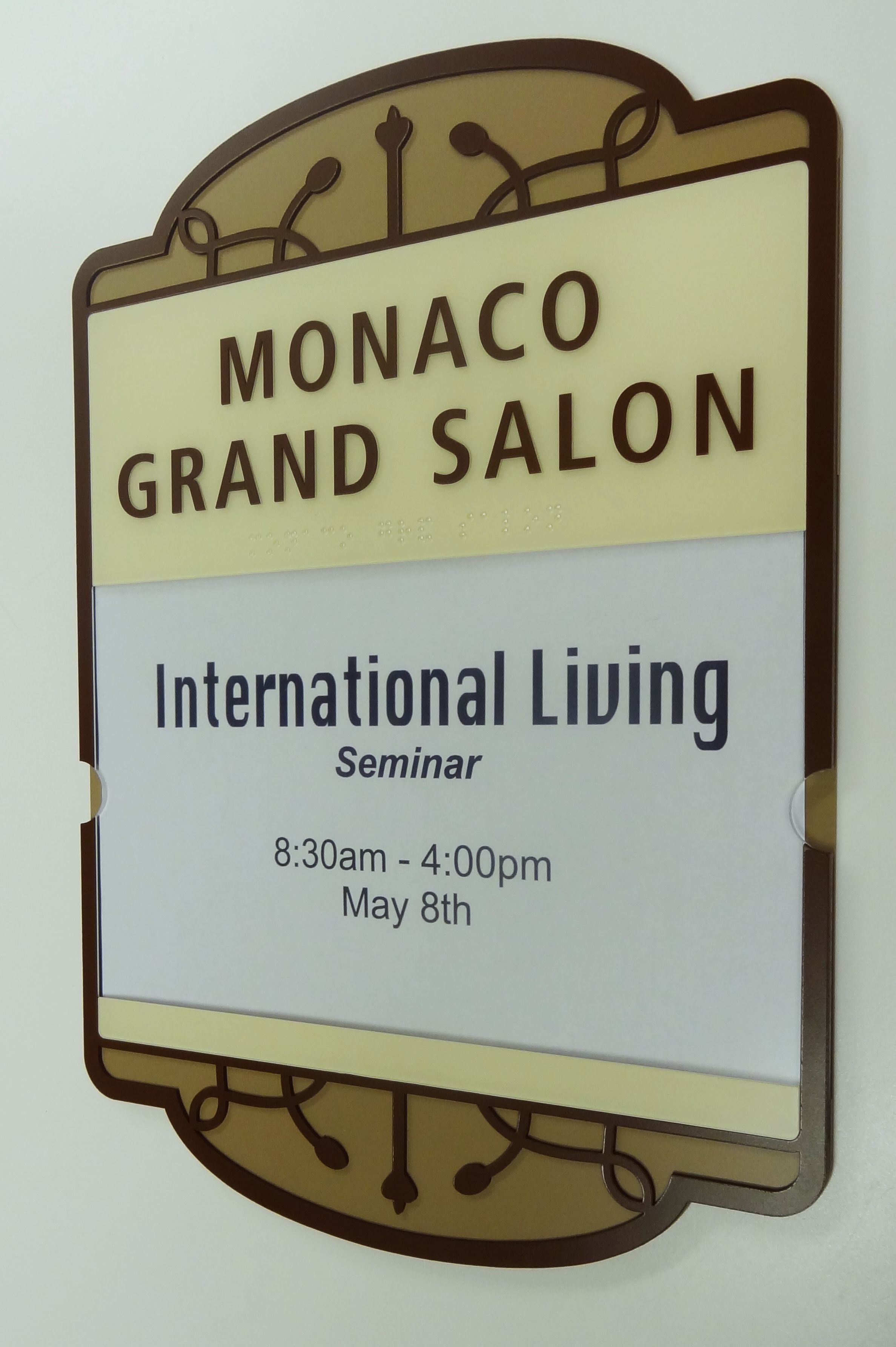A sign on a wall reads "Monaco Grand Salon" at the top, with text underneath announcing an "International Living Seminar" scheduled from 8:30 am to 4:00 pm on May 8th. The sign is in brown and beige tones with a decorative border.