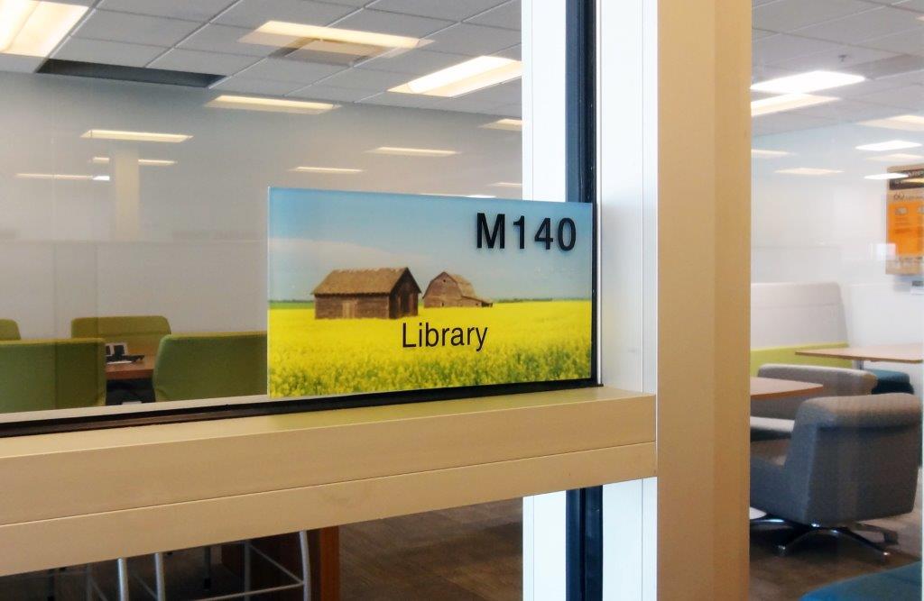 A room with glass walls labeled "M140 Library." The sign includes an image of a rural scene with a barn and field of yellow flowers. Inside the room, there are chairs and tables.