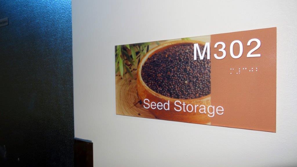 A sign on a wall labeled "M302 Seed Storage" with an image of a bowl filled with dark seeds. Braille text is also present under the room number. The sign has a mix of brown and orange background colors.