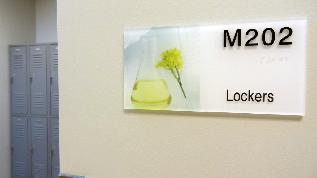 A metal door with a sign reading "M202 Lockers." The sign includes an image of a plant and a beaker with yellow liquid, and braille writing.