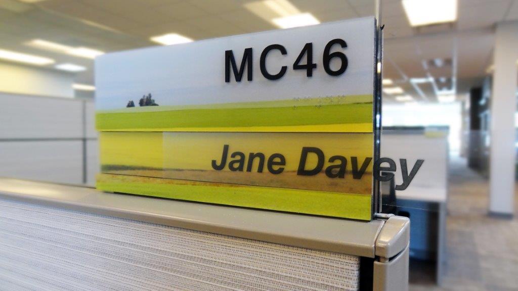 A cubicle nameplate that reads "MC46 Jane Davey" is positioned on the edge of a fabric-covered office cubicle wall in a modern, open-plan office space. The nameplate has an image of a landscape with fields in the background.