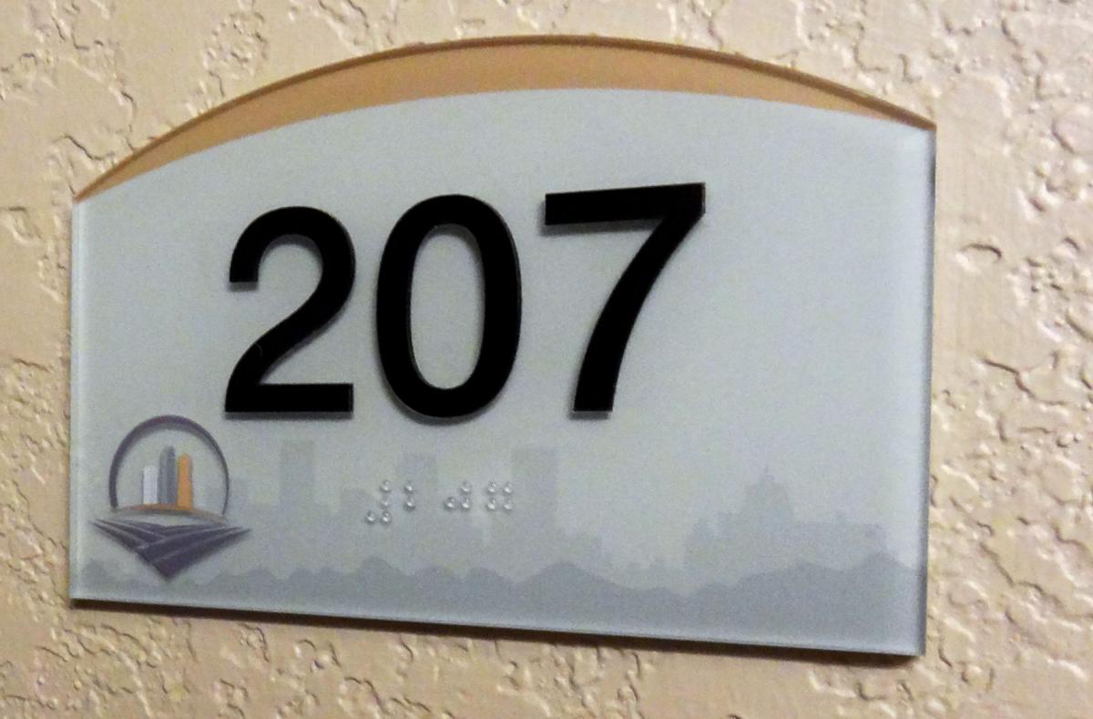 A room sign on a textured beige wall displays the number "207" in large, black print. Below the number is Braille text, and in the bottom left corner, there is a small illustration of a cityscape. The sign is rectangular with a curved top.