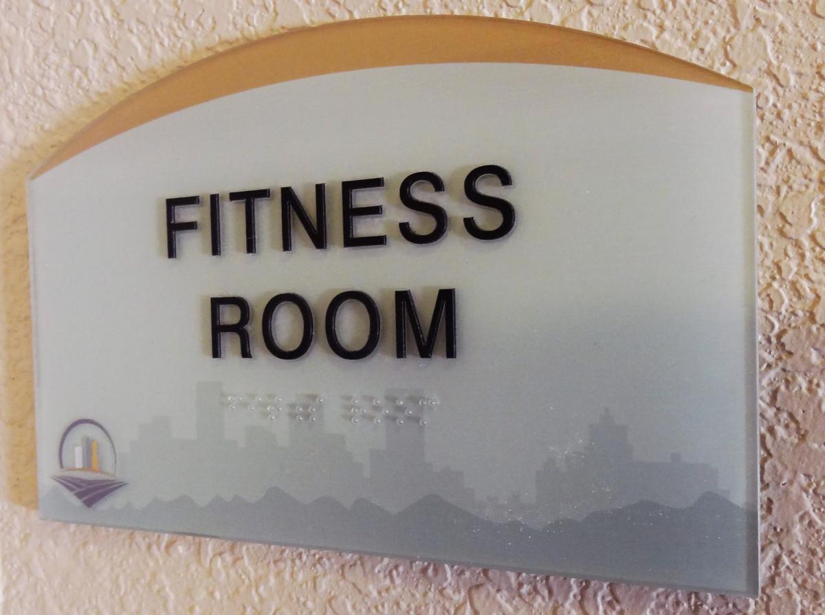 A rectangular sign with a curved top reads "Fitness Room" in bold black letters followed by braille. Below the text, a city skyline graphic is faintly visible along with a colorful logo. The sign is mounted on a textured beige wall.
