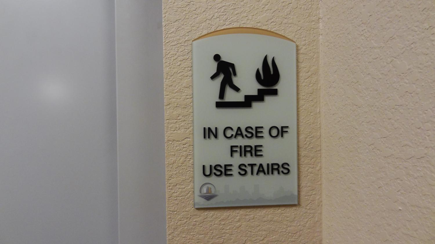 A wall-mounted sign with an icon of a person walking down stairs away from flames. The sign reads: "IN CASE OF FIRE USE STAIRS" in bold capital letters. The background is a textured beige wall adjacent to a doorway with a frosted glass panel.