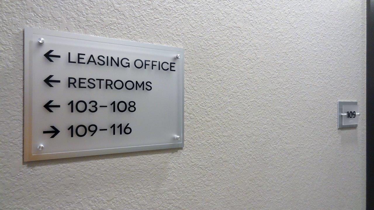 A wall sign with arrows directs to the Leasing Office, Restrooms, units 103-108, and units 109-116. The sign has a frosted glass appearance. A room number sign for room 105 is partially visible on the right wall.