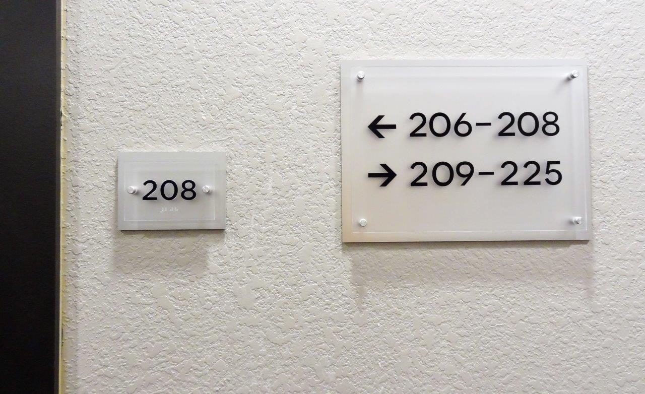 A wall with room number "208" on a small sign. Next to it, a larger sign has arrows pointing left and right. The left arrow indicates rooms 206-208, and the right arrow indicates rooms 209-225. Both signs have black text on a frosted white background.