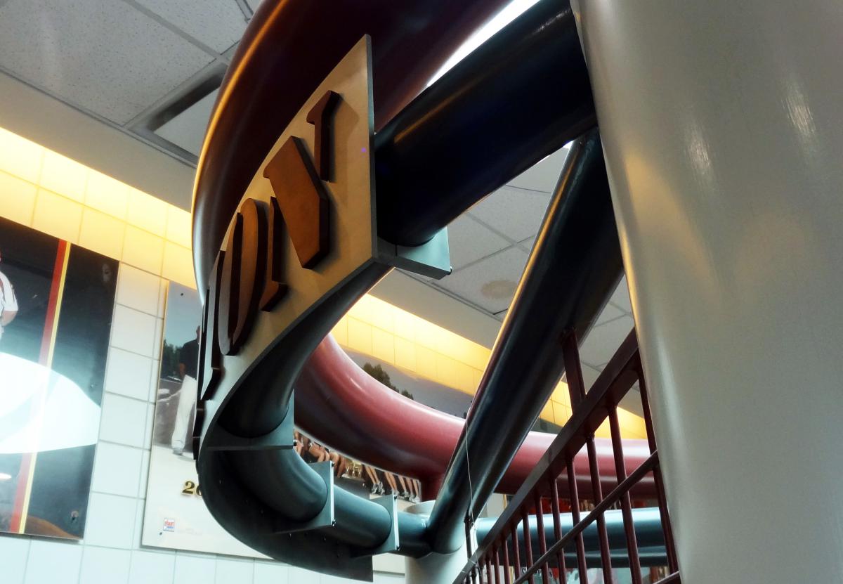 A close-up view of the top section of a large indoor slide, which has the letters "OY" visible on its side. The structure is predominantly dark red and blue, with walls photographed on the left side decorated with images. The ceiling has a grid pattern and light panels.
