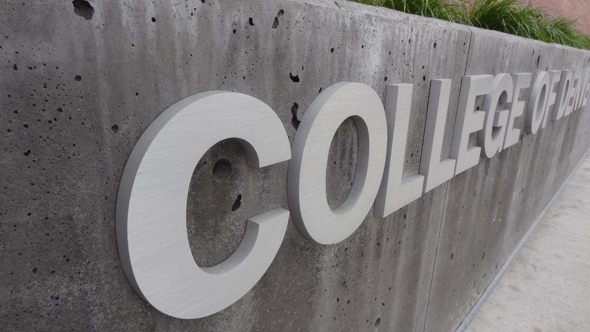 A concrete wall with large white letters reads "Community College of Denver." The wall is located next to a sidewalk and in front of a brick building with tall green plants along the top edge.
