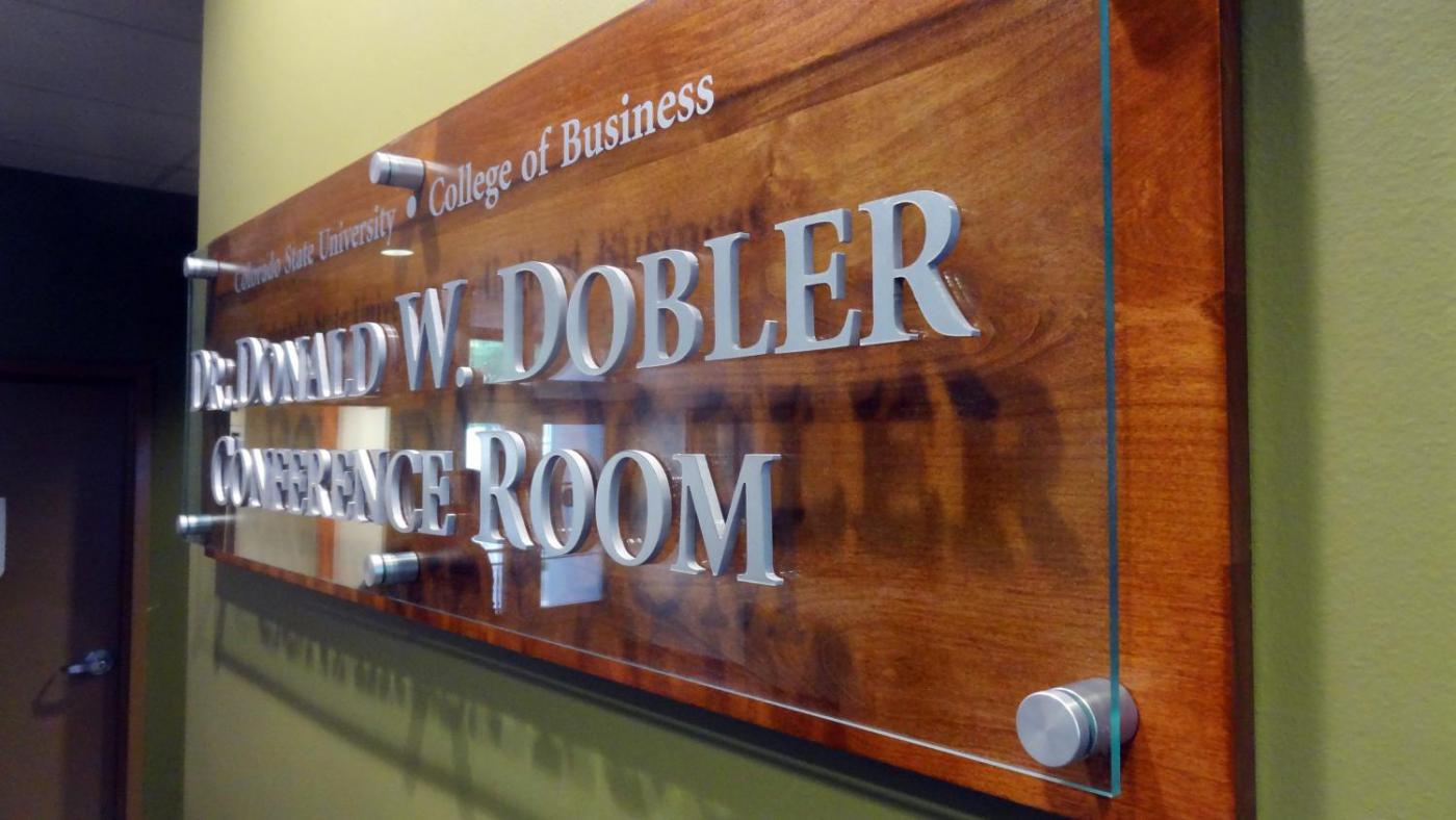A wooden wall plaque with metal letter inlays on glass, affixed to the wood by metal pins. It reads "Dr. Ronald W. Dobler Conference Room" and "College of Business" from Northern State University. The plaque is mounted on a green wall.