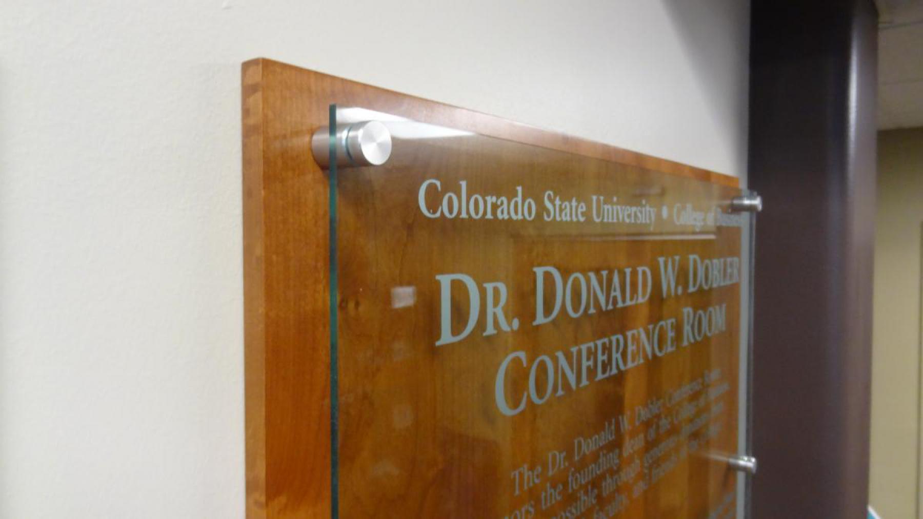 A wooden plaque on the wall reads, "Colorado State University College of Business Dr. Donald W. Dobler Conference Room." The plaque is affixed with four metal caps, which are featured in this closeup.