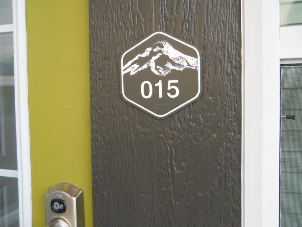A door with an electronic lock has a sign displaying the number "015" in white. The sign is hexagonal with an illustration of a mountain range above the number. The door is dark brown with visible wood grain and is set against a green and white frame.