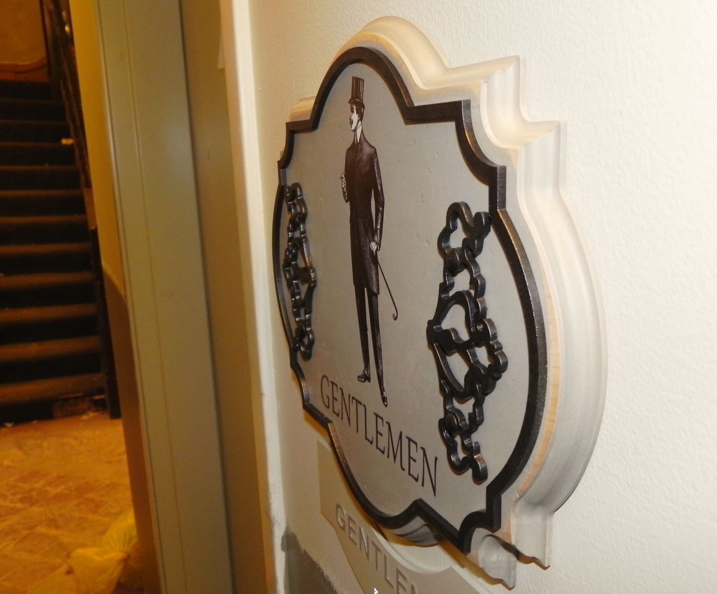 A wall sign reading "Gentlemen" is mounted in a decorative frame featuring an illustration of a man in formal attire, holding a cane and wearing a top hat. The sign is positioned near a staircase, and construction materials are visible in the background.