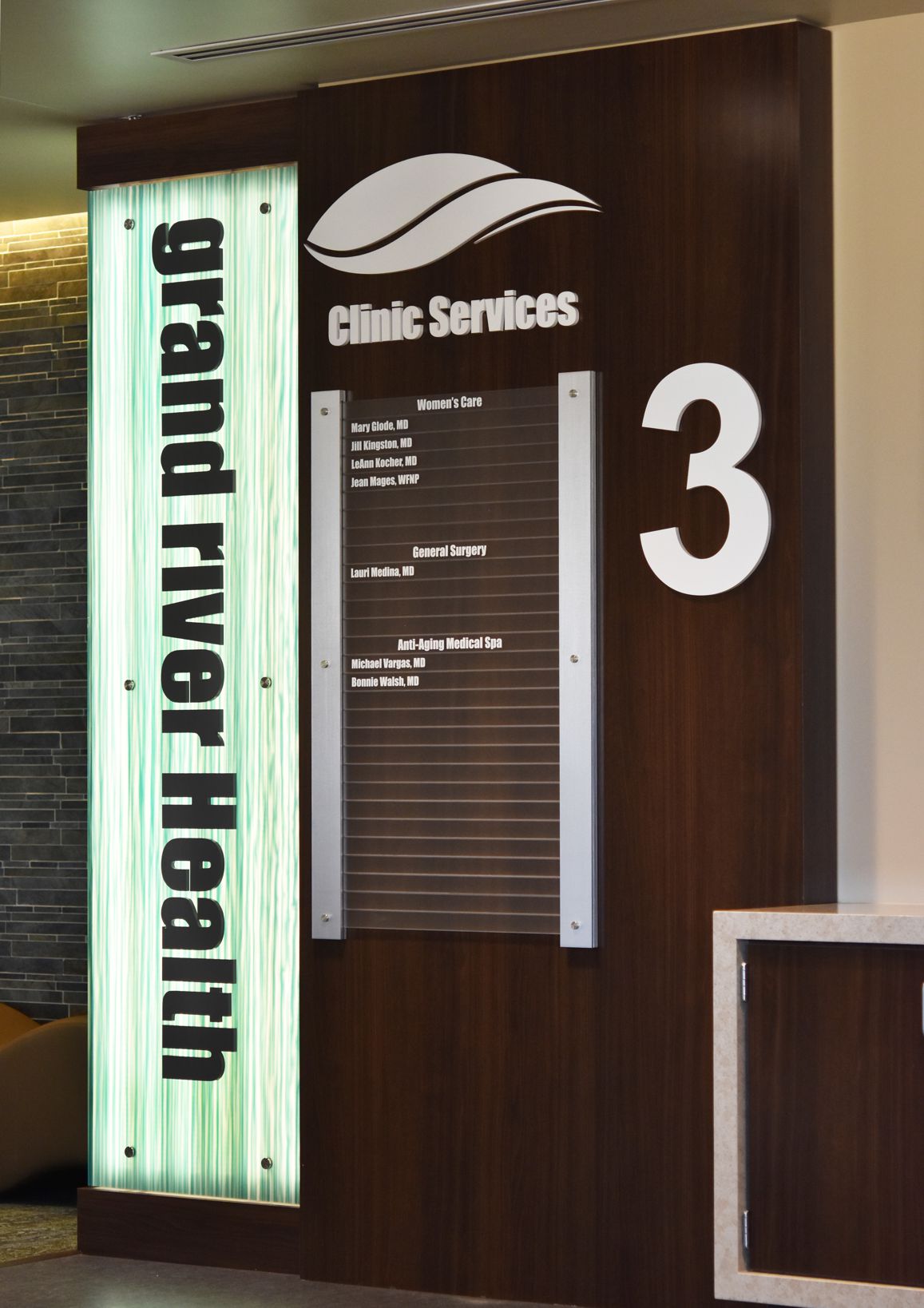 The image shows an illuminated sign for "Grand River Health" next to a directory board for "Clinic Services." The directory lists various departments and room numbers, with a large number "3" to the right of the sign. The decor appears modern and sleek.