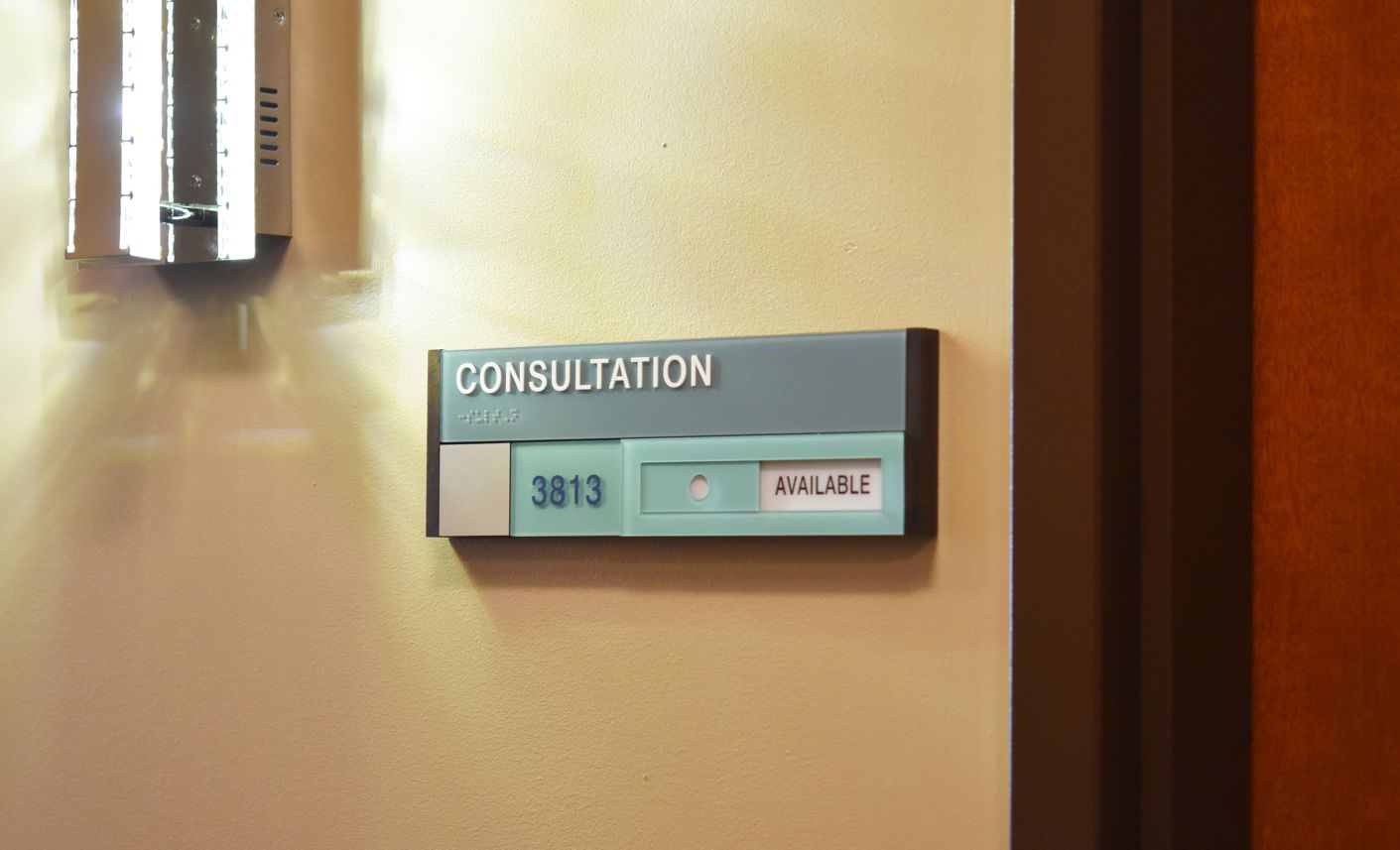 A consultation room door sign reads "CONSULTATION" with room number "3813." The availability indicator shows "AVAILABLE" with a green tab. A portion of a light fixture is visible on the wall next to the sign and door frame.